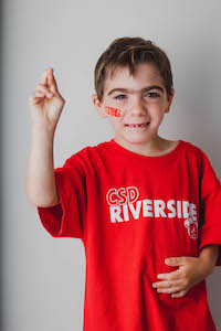 A male young student signing 'R' wearing a red T-shirt with CSD Riverside lettering